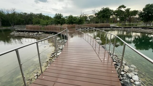 Park boardwalk forward motion over lake, curved wooden path, wet deck, railings, green trees, urban nature, travel recreation.