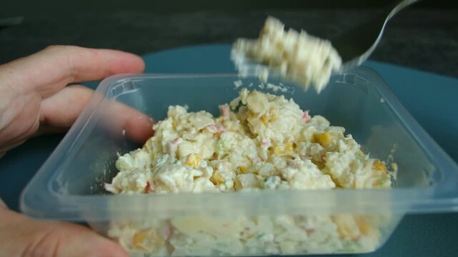 Close up hands holding transparent plastic container eating crab stick salad with metal fork. Convenient but unhealthy meal for quick lunch at office. Person eating crab stick salad, plastic container