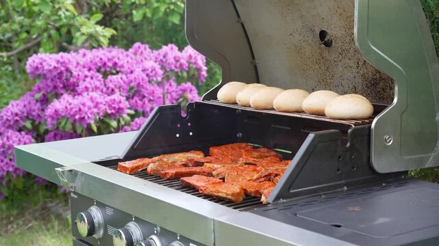 Pork steaks and bread rolls cook on an open garden grill. Purple rhododendron flowers add a colorful summer background.