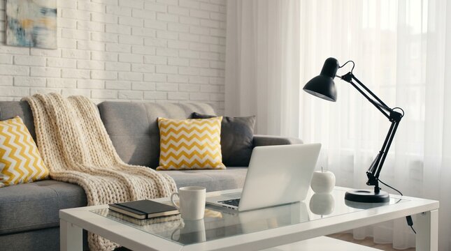 Modern living room interior with laptop on glass coffee table, gray couch with yellow pillows and throw blanket, black desk lamp, and white brick wall background with sheer curtains