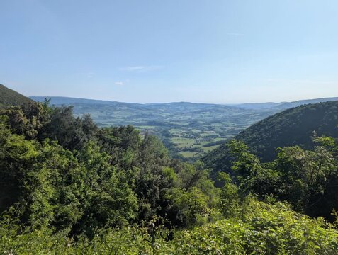 Affaccio panoramico si apre su una vasta pianura coltivata in lontananza, incorniciata da pendii montuosi e alberi rigogliosi in primo piano. 