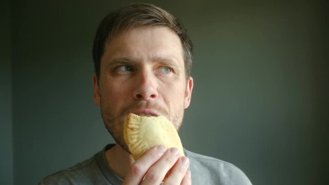 Hungry caucasian man eating delicious empanada with serious expression on face. Person enjoying tasty, savory turnover pastry as quick snack or for lunch break. Caucasian man eating empanada for lunch