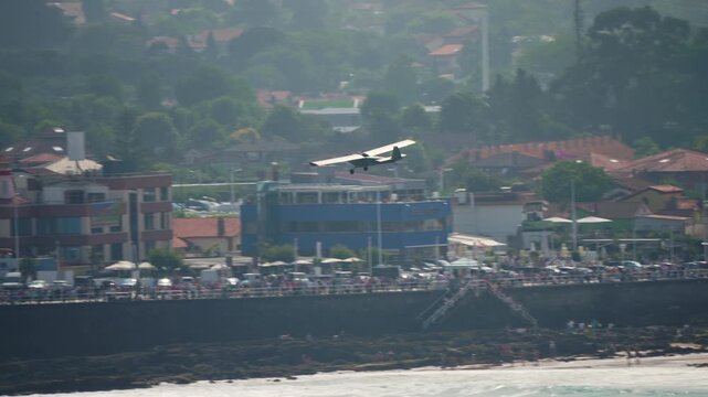 Classic high-wing propeller aircraft in low-level flight along a coastal promenade lined with spectators watching the air display from the waterfront