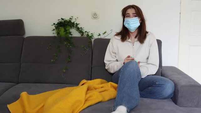 Anxious young woman wearing protective face mask reads alarming news on smartphone and isolates herself at home during possible new pandemic outbreak