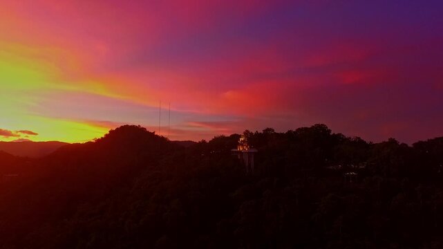 A scenic aerial view of a town nestled among lush green mountains at sunset, with a tower structure perched on the hillside and a vibrant orange sky creating a warm, tranquil atmosphere.