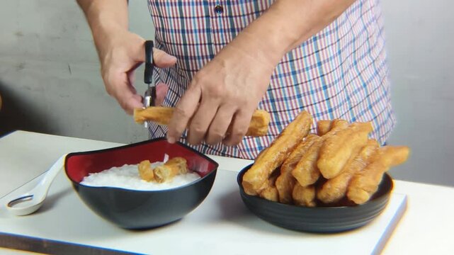 Man's hand cuts fried bread or cakwe using scissors over a bowl of porridge. Eating porridge topped with cakwe is very popular among Chinese people as a breakfast dish.