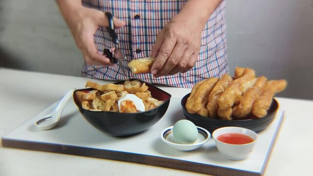 Man's hand cuts fried bread or cakwe using scissors over a bowl of porridge. Eating porridge topped with cakwe and salted egg is very popular among Chinese people as a breakfast dish.