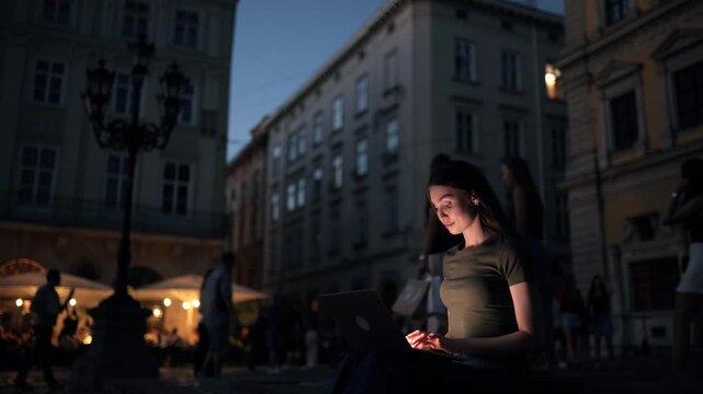 A young brunette woman sits on stone steps in a European city square at night, working on her laptop with a focused and expressive face