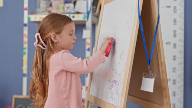 Little caucasian girl erases marker scribbles from a whiteboard using an eraser with her hand in a school building classroom; learning concentration.