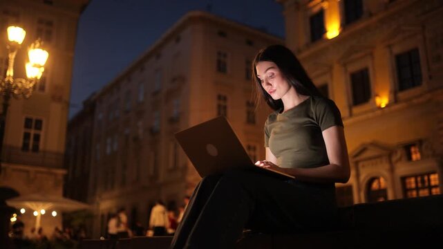 A young brunette woman sits outdoors on stone steps in a historic city square at night, working intently on a laptop