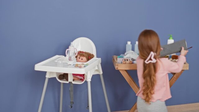 Little girl reaches for baby bottle on wooden tray beside a doll in a highchair, studio; playful nurturing care.