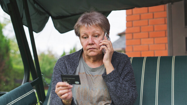 Concerned mature woman talking on smartphone while holding bank card, worried facial expression illustrating online fraud, financial scam, cybersecurity awareness, identity theft concept