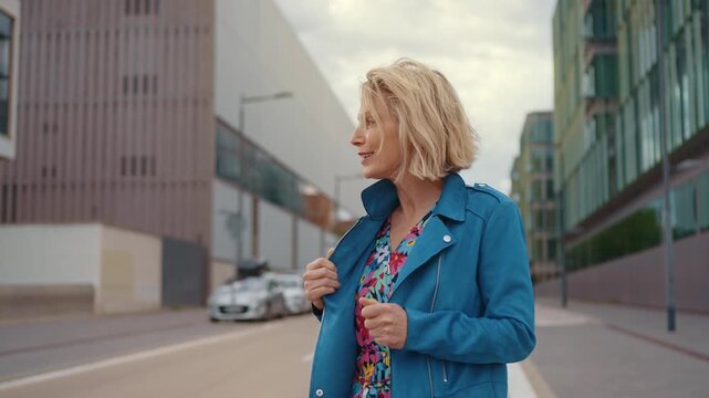 Elegant middle-aged woman with blonde hair enjoying a confident walk through a modern city, smiling with the wind blowing through her hair while wearing a stylish blue jacket and floral dress