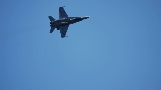 Twin-engine military fighter jet banking steeply in a clear blue sky, aircraft seen from below during aerobatic air display performance