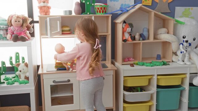 Girl holding a baby doll and placing it on a toy kitchen counter in a preschool building filled with storage bins and stuffed toys; nurturing joy.