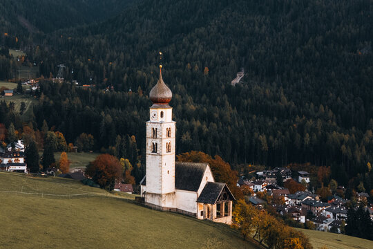 Aerial view of Chiesa di san Valentino with its iconic onion dome tower set against lush green meadows and a dense pine forest in Kastelruth, Trentino-Alto Adige, Italy.