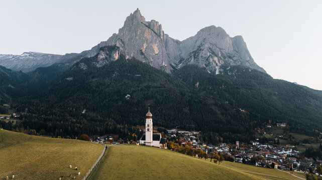 Aerial view of Chiesa di san Valentino with its onion dome tower standing in green meadows against the Schlern mountain peaks in Kastelruth, Trentino-Alto Adige S, Italy.
