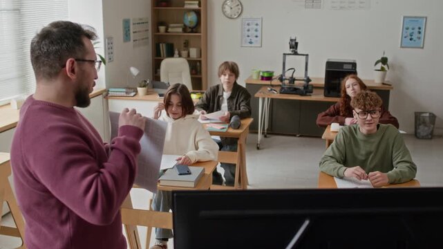 Male teacher holding test paper and explaining topic to students, listening to presentation on screen in modern classroom