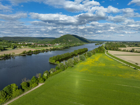 Aerial view of the Danube river winding through lush green fields and the town of Donaustauf under a cloudy blue sky in Donaustauf, Bavaria, Germany.