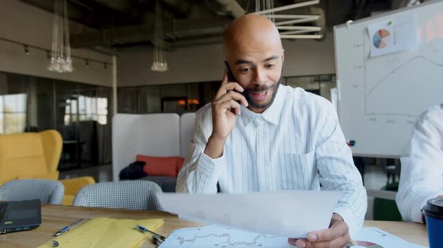 In a contemporary office with ample natural light, a businessman is engaged in an important phone conversation as he attentively reviews various documents spread out before him.