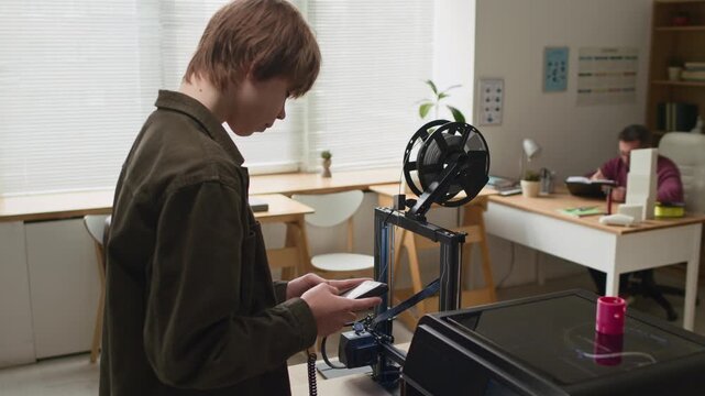 Student intently adjusting print settings on portable control screen in front of 3D printers, while instructor checking students work by window in brightly lit classroom