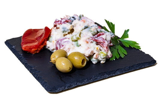 Ensaladilla Rusa Casera, con hoja de perejil, olivas rellenas y pimientos del piquillo sobre una plato de pizarra negro.