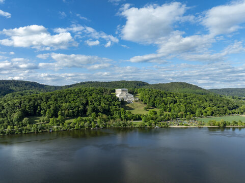 Aerial view of the Walhalla memorial perched on a lush green hill overlooking the Danube river under a bright blue sky with scattered clouds in Donaustauf, Bayern, Germany.