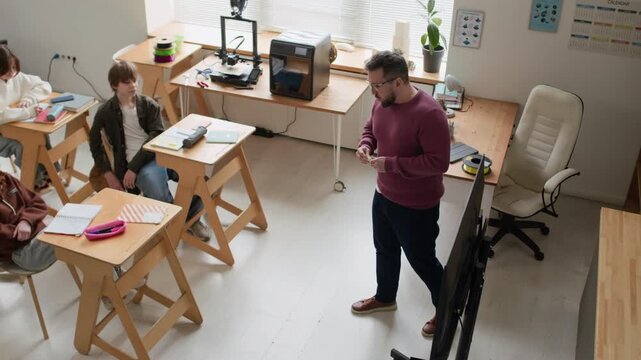 High angle shot of male teacher, holding printed part, explaining its structure to students during 3D modeling class