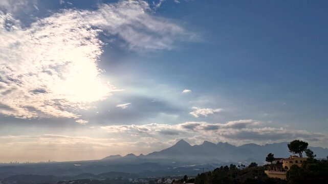 Aerial hyperlapse over Sierra de Bernia, Alicante, Spain. Dramatic crepuscular rays burst from behind fast moving clouds above iconic mountain range with Mediterranean villa below.