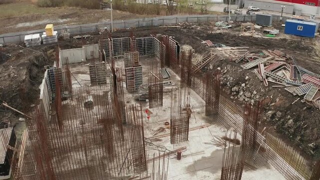 Aerial flyover of a new building foundation under construction. Shows reinforced concrete base, steel rebar cages for columns walls, formwork. Muddy ground construction materials surround site
