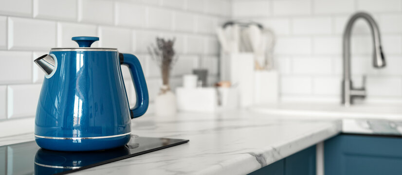 Blue electric kettle on a marble countertop in a modern white kitchen with subway tile backsplash and soft natural light.