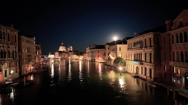 Night time-lapse video of a full moon rising over the Grand Canal in Venice, Italy. Fast moving boats with light trails and illuminated Santa Maria della Salute basilica.