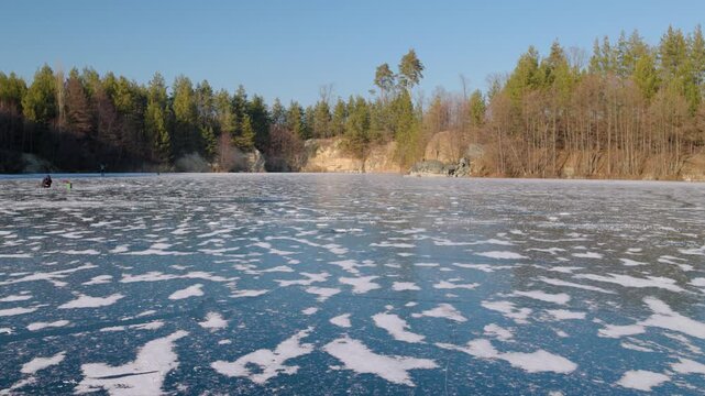Scenic wide shot of a frozen forest lake covered with snow patterns on a sunny winter day. Fishermen are visible on the ice with a beautiful pine forest and clear blue sky in the background.