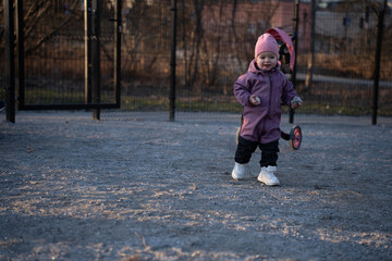 Fototapeta premium toddler in a pink beanie and purple snowsuit walking on gravel near a black metal fence at dusk, clutching small stones beside a parked stroller, warm low light and muted autumn tones, smile, cold