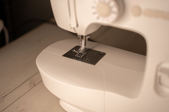 close-up of a modern sewing machine needle and presser foot on a metal throat plate, warm sepia lighting and shallow depth of field over a pale wooden table, evoking a calm minimal domestic craft mood