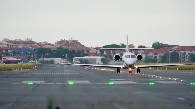 White twin-engine private jet rolling head-on along runway with green threshold lights, harbor boats and coastal buildings visible beyond