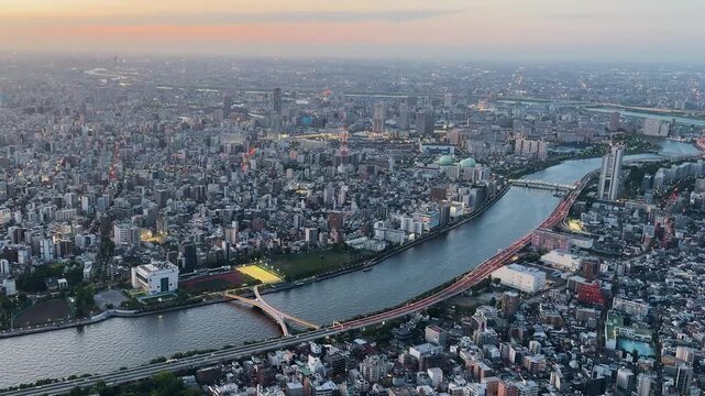Aerial high-angle footage captures a panoramic evening view of Tokyo city from the Tokyo Skytree observation deck. The camera overlooks the winding Sumida River with its iconic bridges. 