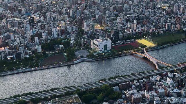 Aerial high-angle footage captures a panoramic evening view of Tokyo city from the Tokyo Skytree observation deck. The camera overlooks the winding Sumida River with its iconic bridges. 