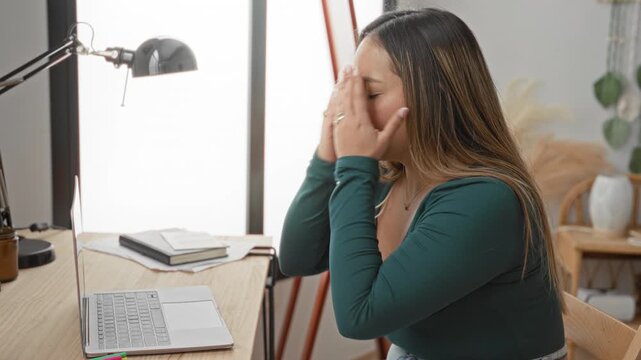 Woman typing on laptop at a desk, hands pressed to face and eyes closed, appearing strained in a studio with lamp and notebook beside her; quiet frustration.