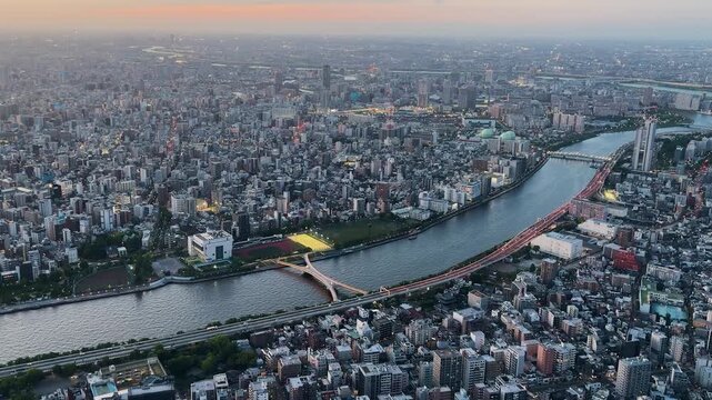 Aerial high-angle footage captures a panoramic evening view of Tokyo city from the Tokyo Skytree observation deck. The camera overlooks the winding Sumida River with its iconic bridges. 
