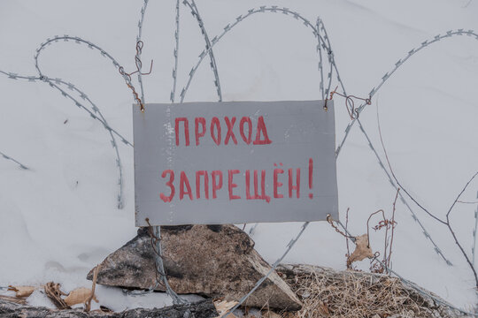 Warning sign in Russian stating "Access Forbidden" attached to barbed wire fence with snow-covered ground and rocks visible in the background