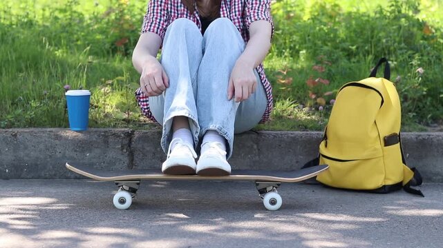 Young girl resting on the side of the road with a skateboard and a yellow backpack. Stylish teenager in light jeans and a plaid shirt sits on the curb next to a skateboard. Youth culture concept