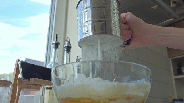 Hand sifts flour from sifter into glass bowl with batter on kitchen counter. Dessert preparation and cooking process concept
