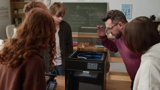 Teacher wearing glasses and burgundy sweater demonstrating inner workings of 3D printer to group of focused and interested students during hands-on class