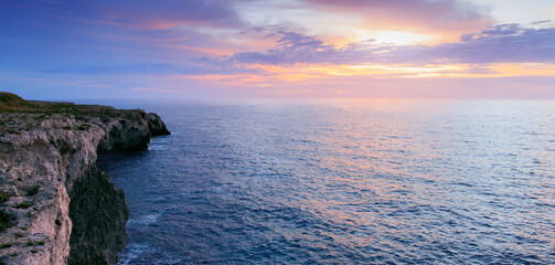 Dramatic ocean cliffs meet a pastel sky at sunset with gentle waves below © Artem Kniaz