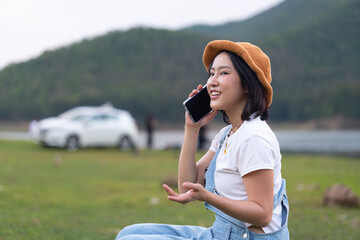 Fototapeta premium Young Asian woman traveler talking on her smartphone while sitting on a rock in a meadow by the lake, with a white car and scenic mountain background during an outdoor road trip.