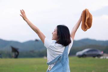 Rear view of a happy young Asian woman traveler raising her arms in the air and holding a hat, celebrating freedom and success in a green meadow with a scenic mountain background © Oil And Den