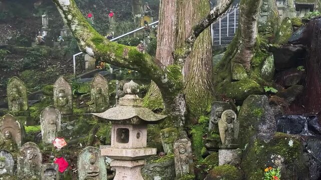 Mossy stone Rakan and Buddhist statues stand on the green hillside of Raizan Sennyoji Daihioin temple grounds in Fukuoka, Japan. A small natural mountain waterfall flows into a stream.