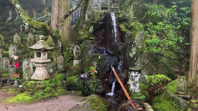 Mossy stone Rakan and Buddhist statues stand on the green hillside of Raizan Sennyoji Daihioin temple grounds in Fukuoka, Japan. A small natural mountain waterfall flows into a stream.