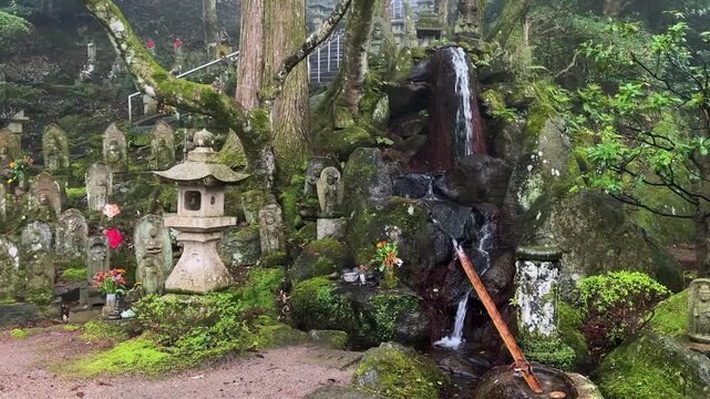 Mossy stone Rakan and Buddhist statues stand on the green hillside of Raizan Sennyoji Daihioin temple grounds in Fukuoka, Japan. A small natural mountain waterfall flows into a stream.